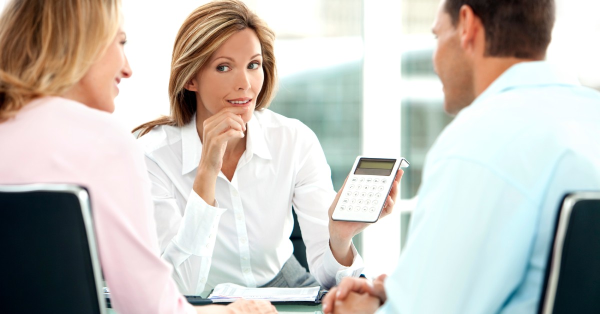 A woman and a couple sitting at a desk. The woman is holding a calculator and showing it to the couple.
